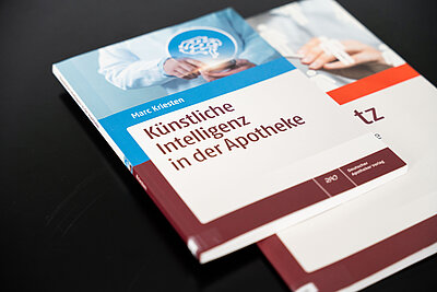 Bücherstapel auf einem Tisch, zu sehen ist das Cover des Buchs "Künstliche Intelligenz in der Apotheke" von Marc Kriesten