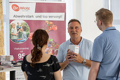 Abgebildet sind drei sich unterhaltende Menschen eine Frau mit schwarzem Kleid und braunen Haaren und ein Mann mit blauem Poloshirt und blonden Haaren sind von hinten zu sehen. Sie unterhalten sich mit einem Man mit hellblauem Poloshirt, silberner Uhr und grauen Haaren. Er hält eine weiße Schachtel in der Hand. Im Hintergrund ist ein roter Banner mit dem Logo "Dr. Wolz" und der Headline "Abwehrstark und top versorgt!"