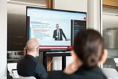 Gruppe von Menschen, die auf einem TV-Monitor den Vorträgen zuschauen