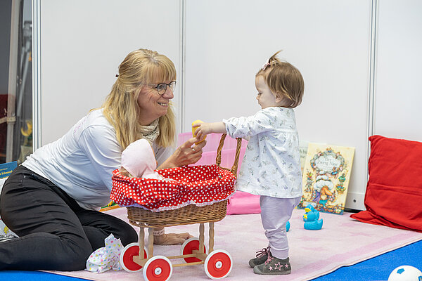 Auf dem Foto ist eine Frau zu sehen, die ein Kleinkind betreut und mit ihm spielt. Zwischen beiden steht ein Puppenwagen.