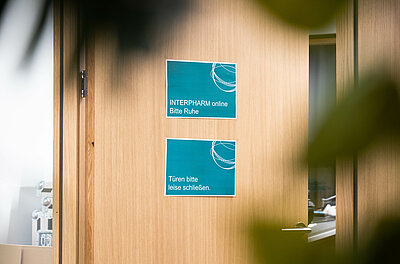 Tür mit türkisnen Schildern mit der Aufschrift "INTERPHARM online, bitte Ruhe. Türen bitte leise schließen." 