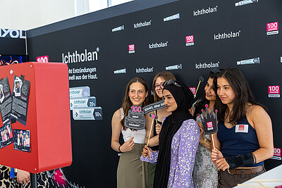 Eine Gruppe von fünf jungen Frauen steht vor einer schwarzen Logowand von "Ichtholan". Vor Ihnen eine rote Fotobox. Sieh halte verschiedene Pappfiguren in der Hand, darunter eine schwarze Torte mit einer 100 in rosa und eine schwarze Krone ebenfalls mit einer rosa 100