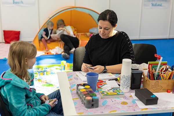 Auf dem Foto ist eine Frau zu sehen, die ein Kind betreut und mit ihm spielt, auf dem Tisch sind Wassermalfarben zu sehen
