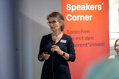 Auf dem Bild ist eine referierende Frau mit schwarzem Kleid, Brille und halblangen grauen Haaren zu sehen. Sie hält in der Hand ein Mikrofon und seht vor einem roten Banner mit der Aufschrift Speakers Corner – Sprechen sie mit den Referen*innen
