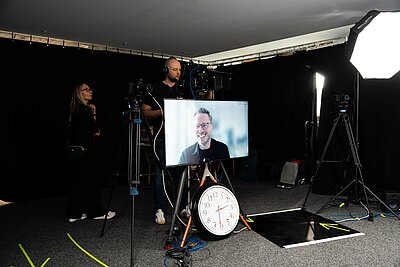 Blick ins Studio, auf einem TV-Monitor ist Referent Chris Günter zu sehen, unter dem Monitor eine große Uhr