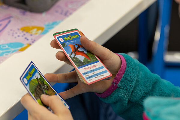 Auf dem Foto sind Kinderhände zu sehen, die Spielkarten halten. Auf einer Karte ist ein Flugdinosaurier abgebildet.