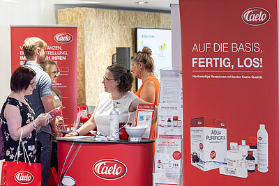 Zu sehen ist ein roter Messestand der Firma Caelo. Im Vordergrund ein Counter mit einer Frau in weißen Top, die einer interessierten Kundin in dunkelblauem Kleid zugewandt ist. Rechts im Bild ist ein rotes Banner mit verschiedenen Produkten dem Caelo-Logo und der Headline "AUF DIE BASIS, FERTIG, LOS!"