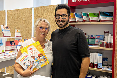 Auf dem Foto sind zwei Personen, die in die Kamera lachen. Ein Mann mit schwarzem T-Shirt, schwarzem Haar und Bart und schwarzer Brille. Neben ihm eine blinde Frau mit Brille und weißem T-Shirt. Sie hält lächelnd zwei Bücher in die Kamera. Das eine Buch ist gelb und heißt "Sag mal aaah!", das andere Buch ist weiß und heißt "Kompass Kindermedizin"