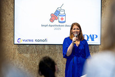 Auf dem Bild ist eine referierende Frau mit blauem Kleid, mittellangen rotblonden Haaren zu sehen. Sie hält in der Hand ein Mikrofon und seht vor einem weißen Bildschirm mit der Aufschrift Impf-Apotheken-Award 2025 und einer Grafik mit einer Spritze und einer Dose mit rotem Kreuz