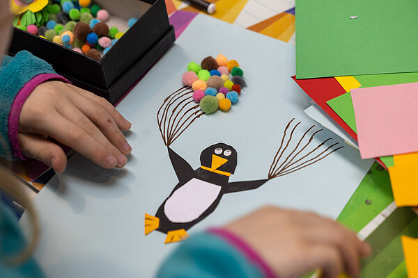 Auf dem Foto sind Kinderhände zu sehen, die ein Bild malen. Auf dem Bild ist ein Pinguin mit Luftballons aus Filz zu sehen