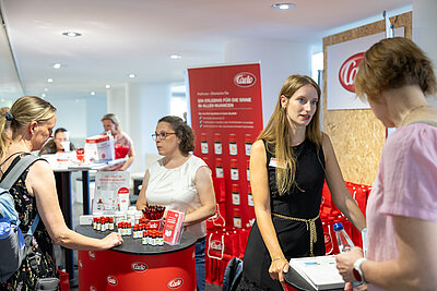 Auf dem Bild ist ein kleiner Messestand zu sehen. Im Vordergrund ein roter Counter mit kleinen Fläschchen im Hintergrund eine rote Werbewand. Auf dem Foto unterhalten sich vier Personen.