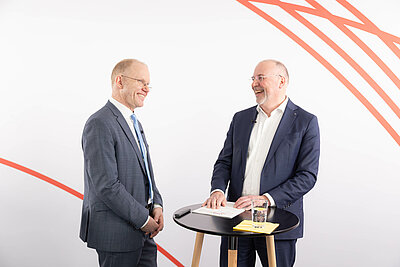 Dr. Hubert Ortner im Gespräch mit Johannes Bauernfeind. Beide stehen vor weißem Hintergrund an einem Stehtisch und lachen.