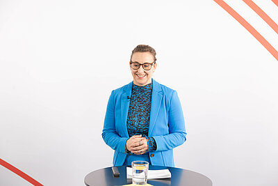 Melanie Dolfen steht im blauen Hosenanzug und gefalteten Händen an einem Stehtisch vor weißem Hintergrund