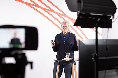 Prof. Dr. Reinhard Herzog ist in der Bildmitte zu sehen, Blick nach unten, gestikulierend. Flankiert ist er im Vordergrund von Kamera und Lichtequipment.