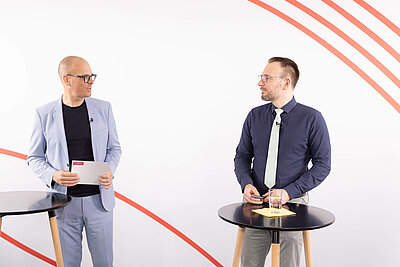 Dr. Benjamin Wessinger und Dr. Christian Knobloch stehen nebeneinander und unterhalten sich. Beide haben jeweils einen Stehtisch vor sich.
