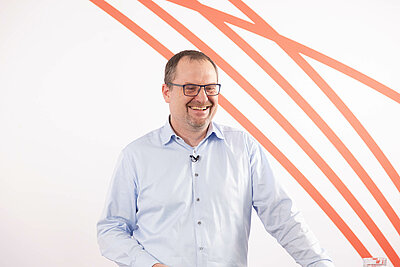 Dr. Morton Douglas in hellblauem Hemd lachend vor weißem Hintergrund mit rotem Streifenmuster