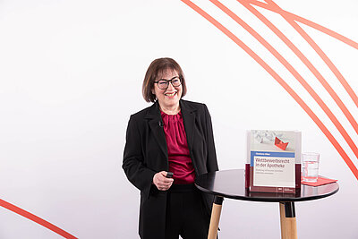 Christiane Köber steht lachend neben einem Stehtisch, Auf dem Tisch steht das Buch Wettbewerbsrecht in der Apotheke