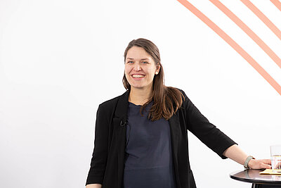 Heike Gnekow schaut lachend nach links an der Kamera vorbei. Sie steht sich mit einer Hand abstützend an einem Stehtisch.