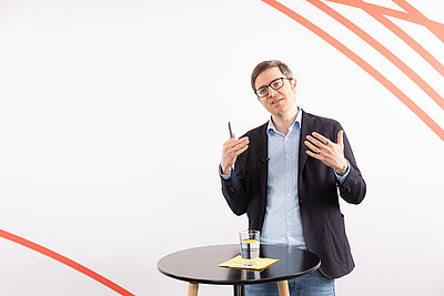 Dr. Björn Schittenhelm steht referierend und mit den Händen gestikulierend hinter einem Stehtisch vor weißem Hintergrund.
