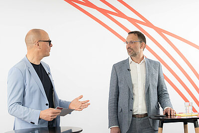 Dr. Benjamin Wessinger und Lorenz Weiler stehen nebeneinander und unterhalten sich. Beide haben jeweils einen Stehtisch vor sich.