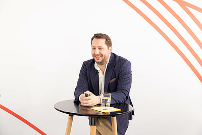 Dr. Moritz Wollring in dunkelblauem Sacko hat sich mit seinen Armen auf einem Stehtisch abgestützt und lacht.