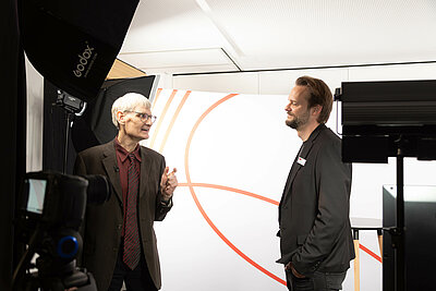 Prof. Dr. Reinhard Herzog ist im Gespräch mit Jan Schüle zu sehen.  zu sehen. Flankiert sind beide von Kamera- und Lichtequipment.